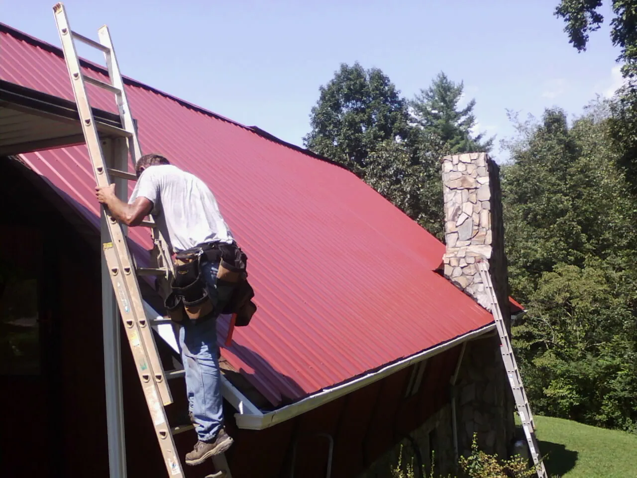 Roofer on a ladder beside a red metal roof and stone chimney — Soffit & Fascia Repair by Roofing Brothers in Fanwood